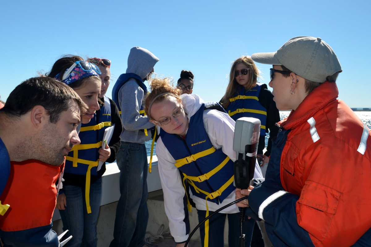 A group of students in life jackets look at a small gray instrument that an instructor holds up