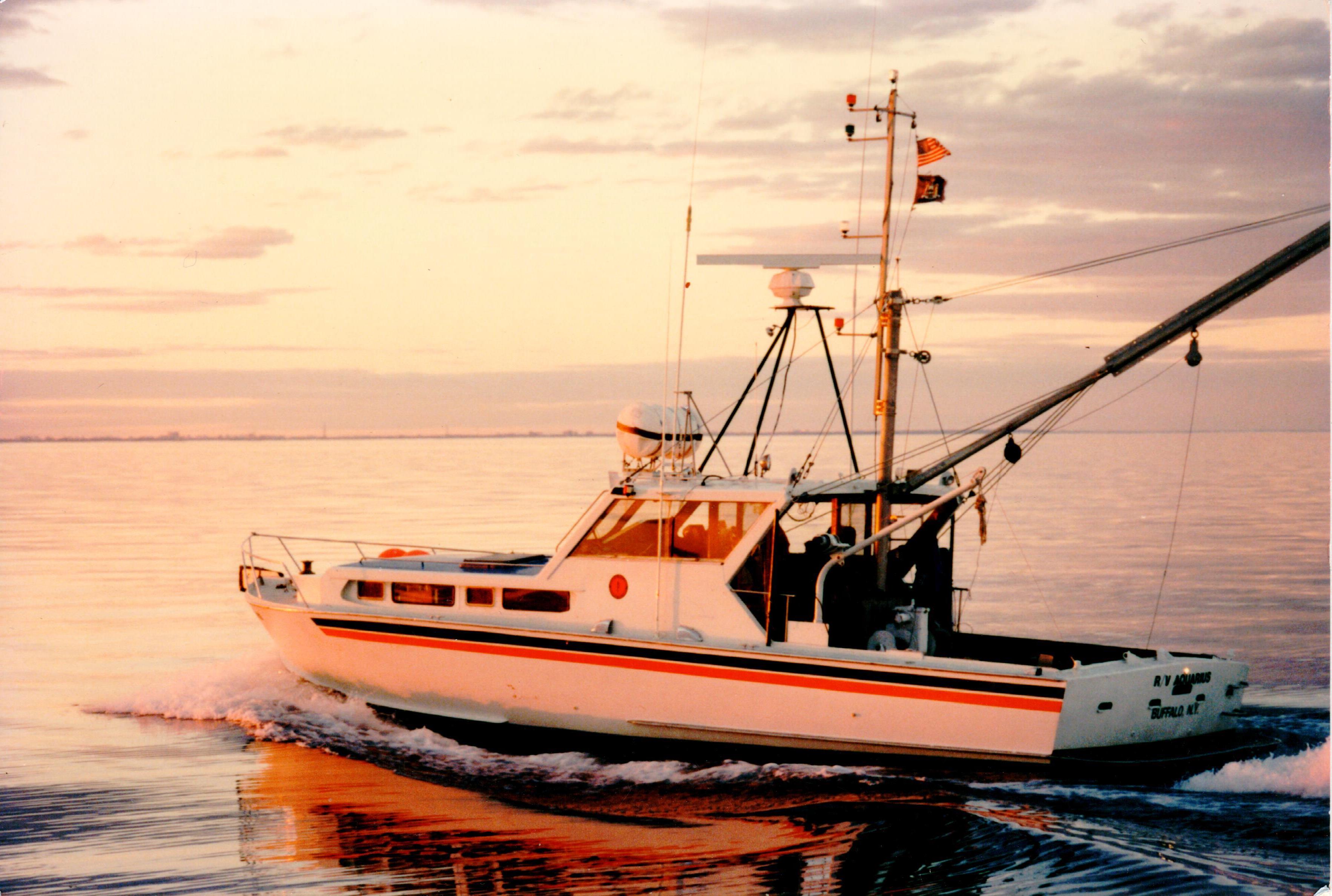Color picture of a boat at sunset, a white boat with an orange & black stripe. A long boom arm extends from the back