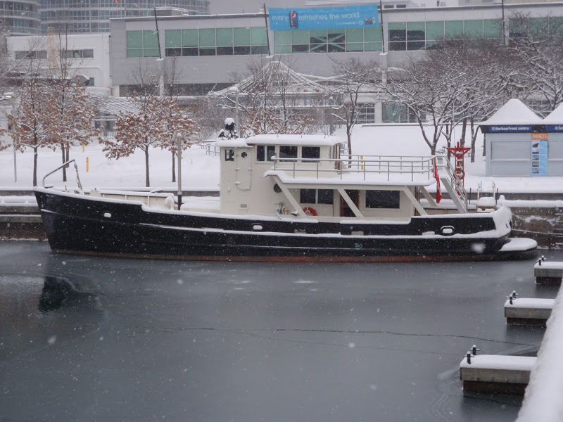 A color photo of a black & white boat docked in icy water in front of some buildings in a snowy city