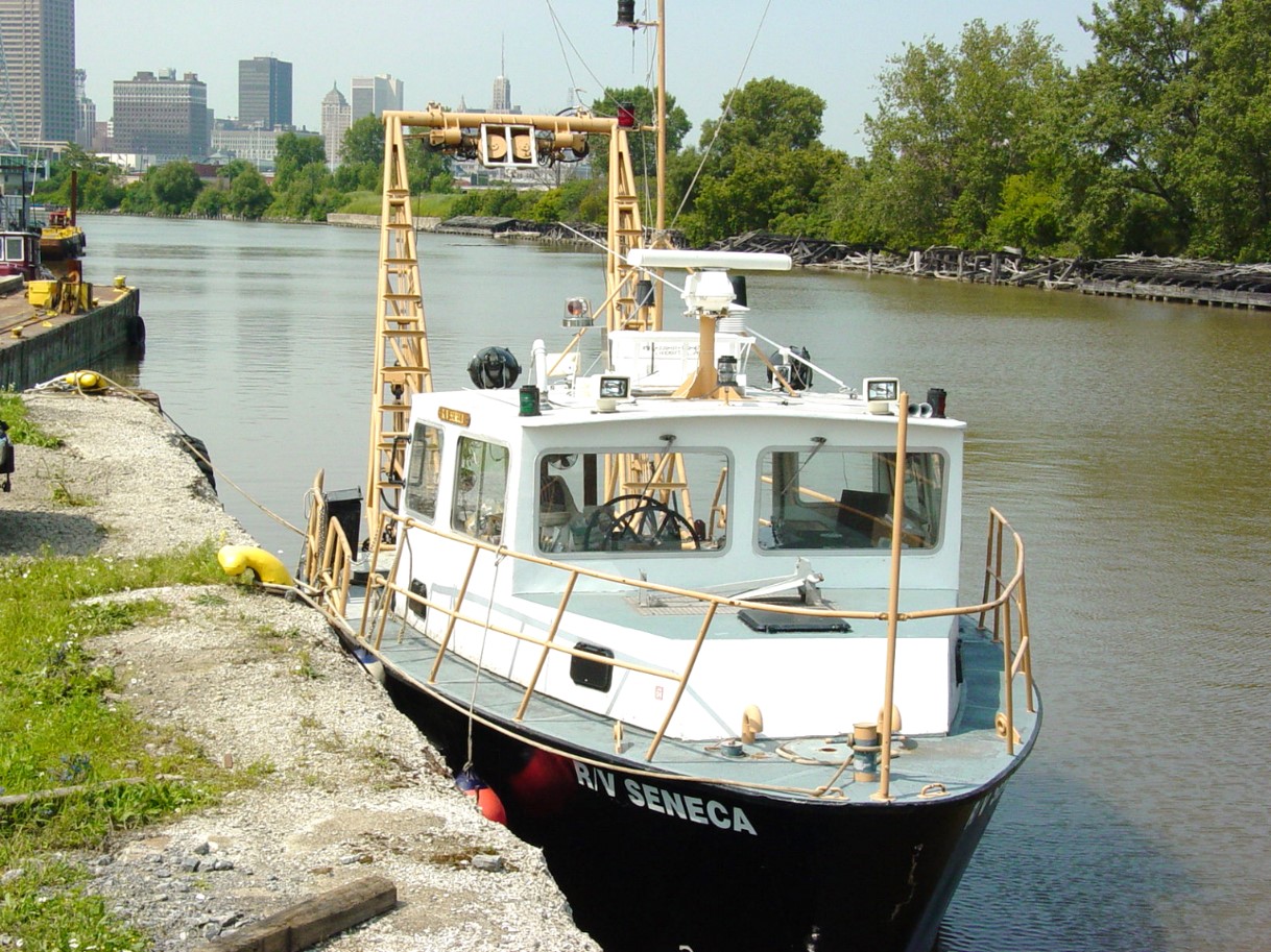A black & white boat, docked in a canal in front of the Buffalo skyline. It has a yellow A-frame boom