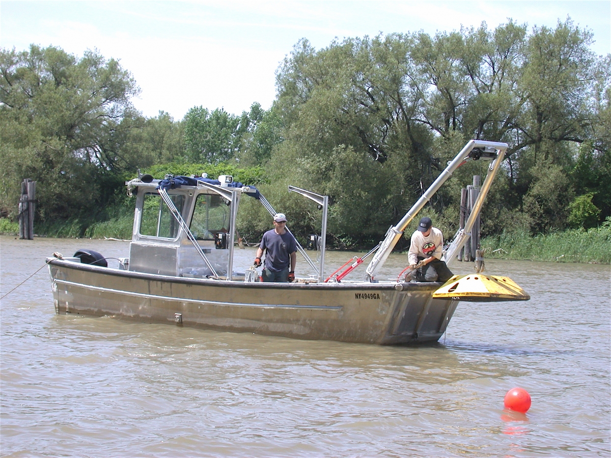 two people on a silver boat in a river use a boom to life a large yellow piece of equipment