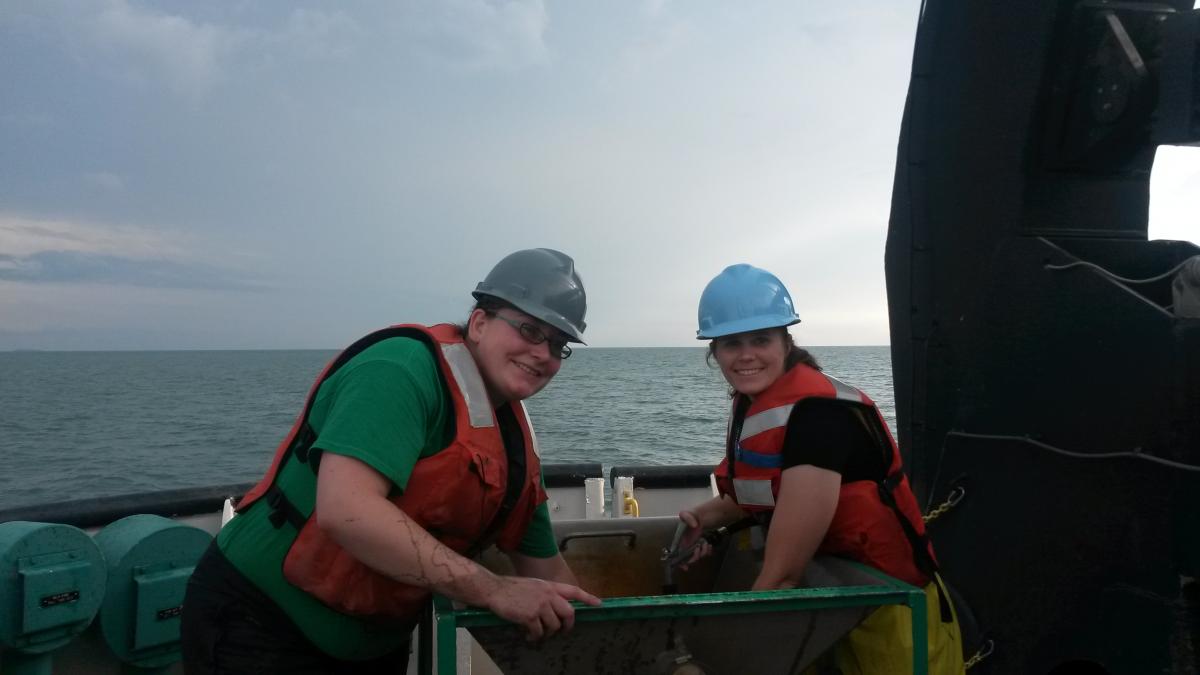 Two people wearing hard hats and life jackets stand at a large basin on the deck of a boat