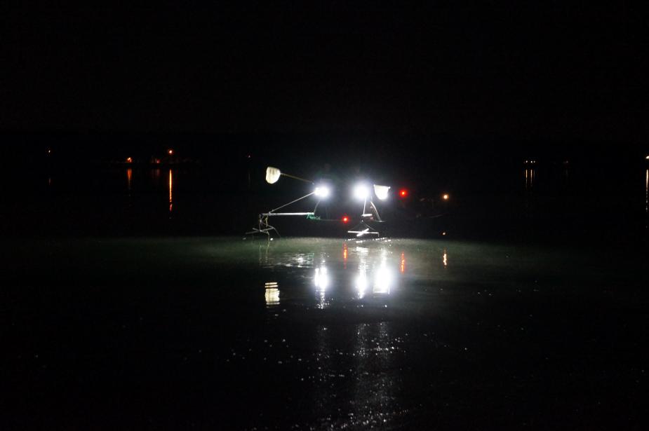 nighttime photo of a boat with bright headlights and two booms extended over the water, and two people with nets