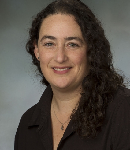 portrait of Andrea Locke, a light-skinned person with mid-chest length curly dark hair wearing a black collared shirt and a small pendant on a necklace