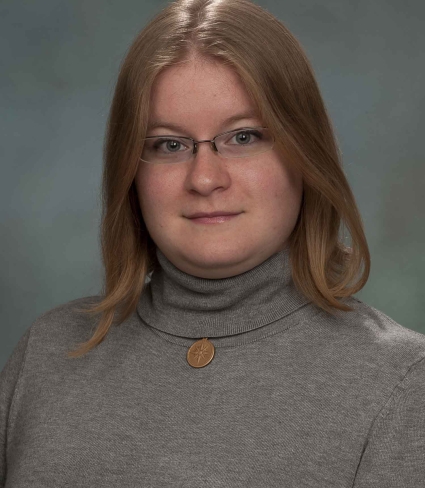 portrait of Brianne Tulumello, a light-skinned person with shoulder-length dark blonde hair, grey eyes, and glasses, wearing a grey turtleneck with a short necklace with a round pendant