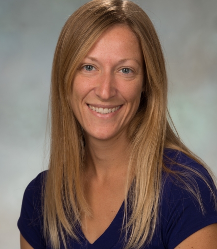 portrait of Susan Dickinson, a tanned light-skinned person with grey eyes and mid-chest length blonde hair, wearing a royal blue V-neck t-shirt
