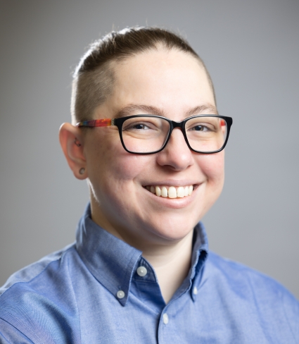 a person a pulled-back undercut with shaved sides smiles, wearing black-framed glasses with rainbow-colored arms and a blue button-up shirt
