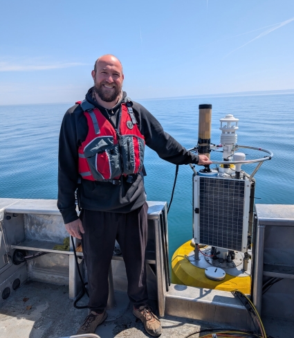A balding person with a beard stands on the deck of a boat holding the top of an observational buoy on a clear, calm day