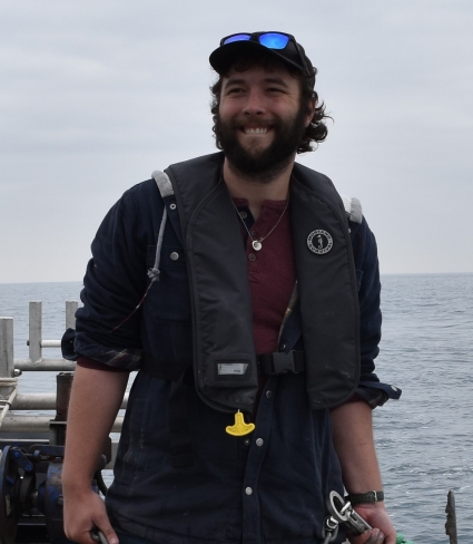 a person with a beard, baseball hat, and sunglasses holds an anchor while working on a boat on an overcast day