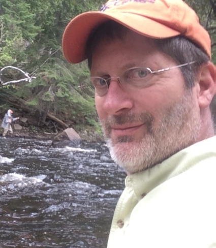 A person with a baseball hat, rounded glasses, and a graying beard by a river in a forest