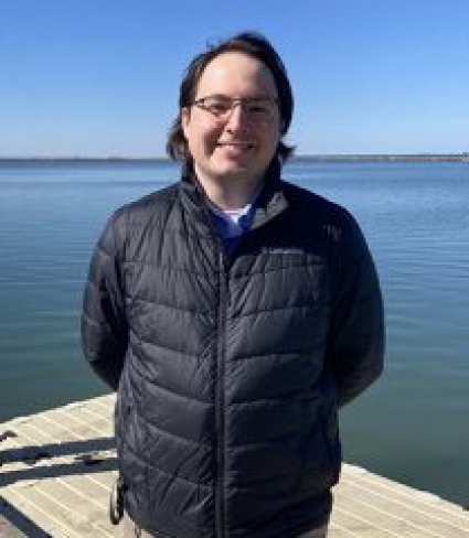 a smiling person with glasses wearing a jacket stands by a dock at the water