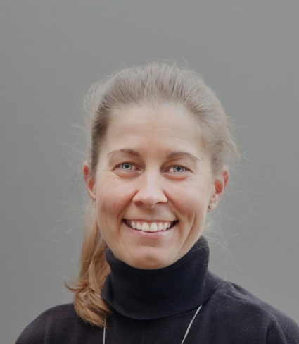 portrait of Rachel Taylor, a light-skinned person with pale blue eyes, a wide smile, light hair pulled back into a shoulder-length pony-tail, wearing a black turtleneck and a necklace