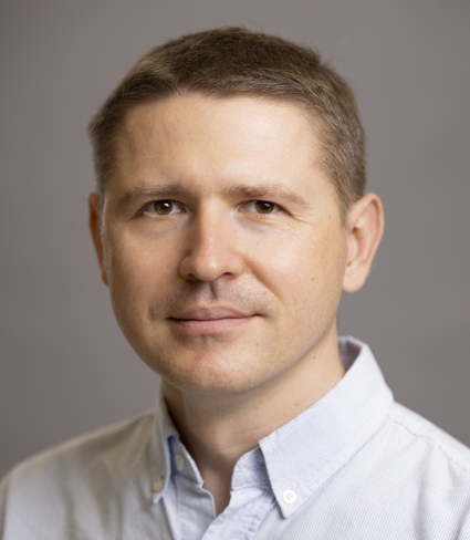 A light-skinned man with short brown hair wearing a light blue striped collared shirt.