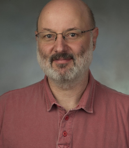 portrait of Thomas Hahn, a light-skinned person that is bald with a graying beard and darker moustache, wearing glasses and a light red polo shirt