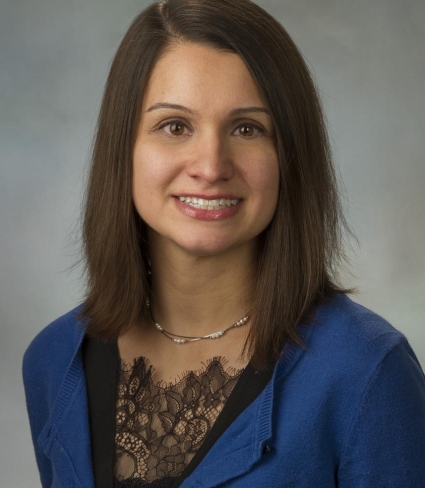 portrait of Brittany Hernon, a tan-skinned person with brown eyes and shoulder-length brown hair parted on one side, wearing a black shirt with a lace embellishment and a blue sweater, and a short necklace with pearlescent beads