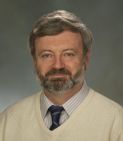 portrait of Alexander Karatayev, a light-skinned person with short graying hair and beard wearing a cream sweater over a striped collared shirt and blue striped tie