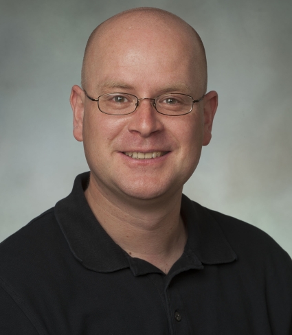 portrait of Knut Mehler, a light-skinned bald person with glasses wearing a black polo shirt