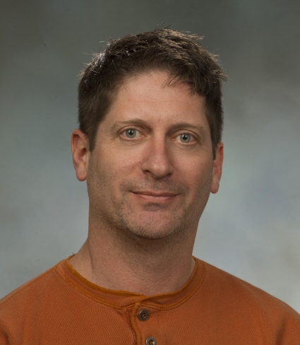 portrait of Christopher Pennuto, a light-skinned person with grey eyes and cropped brown hair wearing an orange shirt with three buttons at the collar