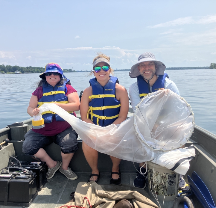 Three people smile and sit on the deck of a small boat on a sunny day, holding a conical net across all of their laps