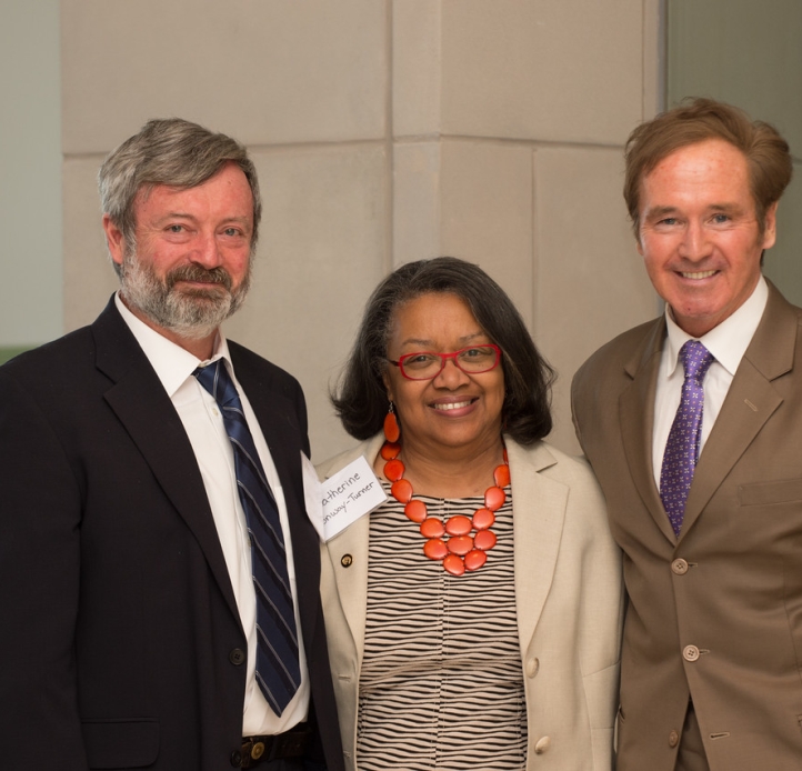 Two people wearing suits and ties stand on either side of a person wearing a suit jacket over a blouse with a chunky necklace made of large orange circles at a reception