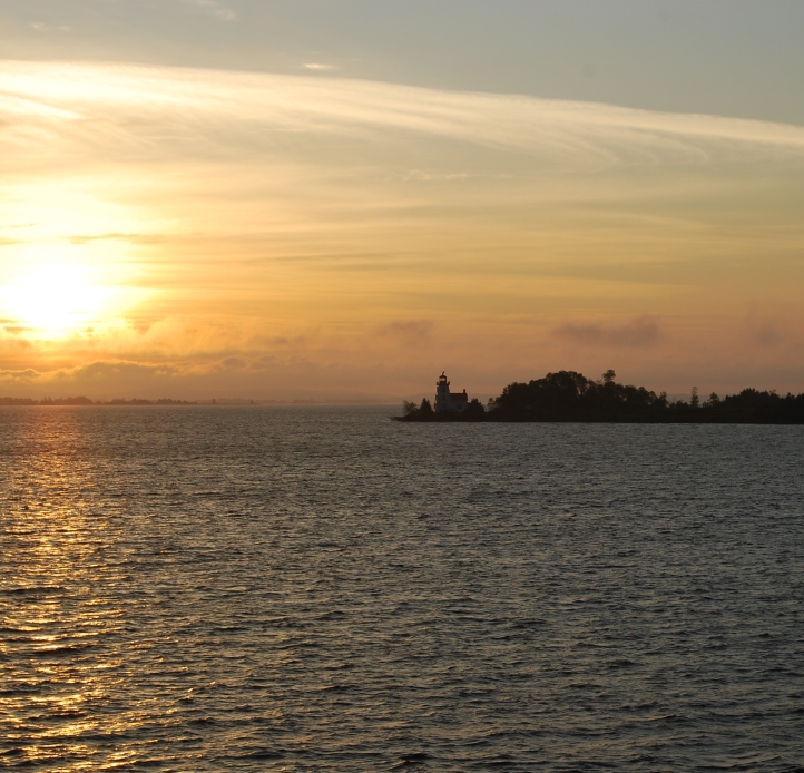 Sun refracting through wispy clouds in the morning above a spit of land with a lighthouse in a large lake.