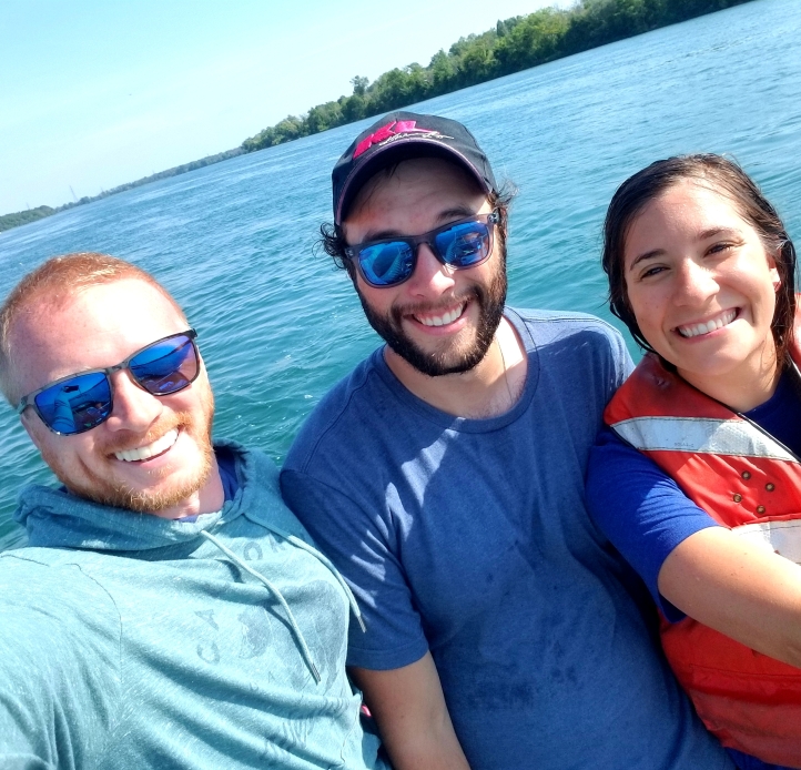Three smiling people stand in front of the water. Two men wearing sunglasses and a woman in a life jacket