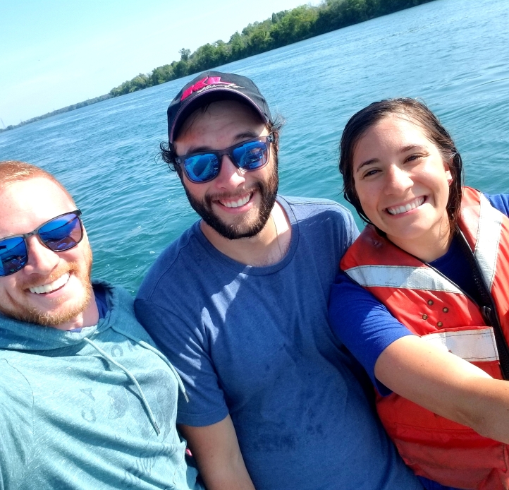 Three smiling people stand in front of the water. One is wearing a life jacket and the other two have sunglasses.
