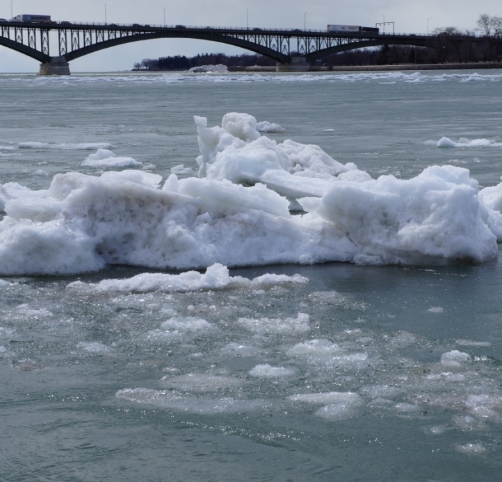 A large chunk of ice in an icy river. There is a bridge in the distance.