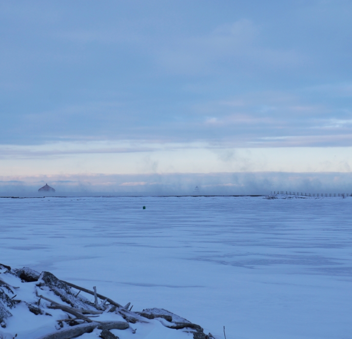 Snow and ice cover a body of water. A breakwall contains the scene, with fog rising from just beyond the breakwall.