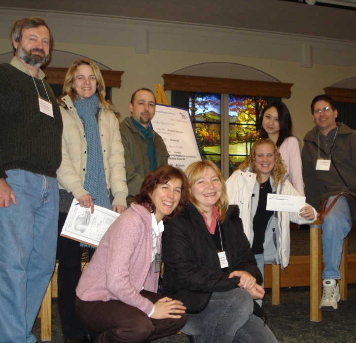 Eight students, researchers, and professors gather to take a picture at a conference by a sign with a schedule on an easel