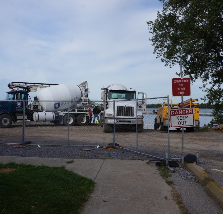 A fence with a sign "DANGER KEEP OUT" blocks a construction zone with two cement trucks and some other equipment. There is a body of water behind the construction zone.