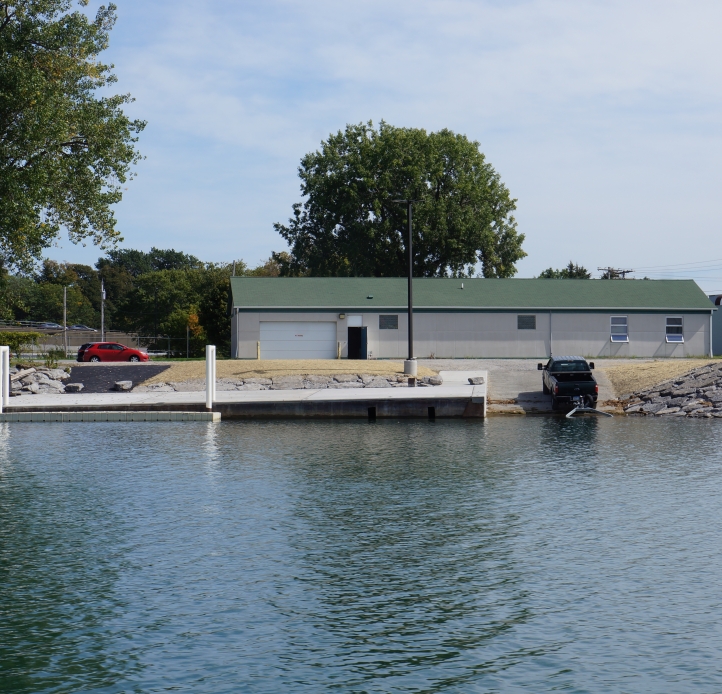 A one-story building with a dock and boat launch next to the water. A truck is backed into the water with a trailer.