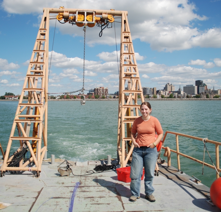 A person stands with their hands in their pockets at the back of a buoy tender boat with a city skyline and fluffy clouds in the background