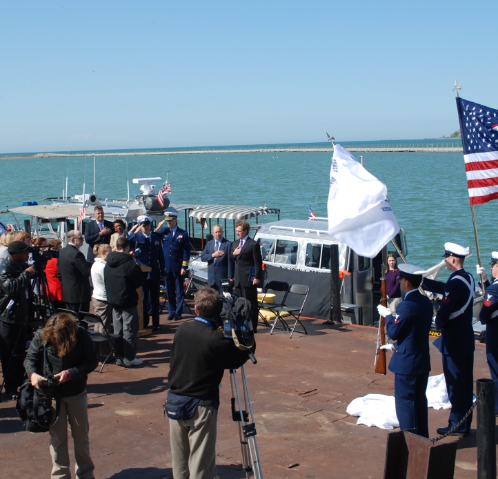 A large group of people assembled for a solemn event on a dock by the water for the christening of a boat. Military color guards furl flags and rifles, and photographers and videographers record as attendees salute or put hands over their hearts.