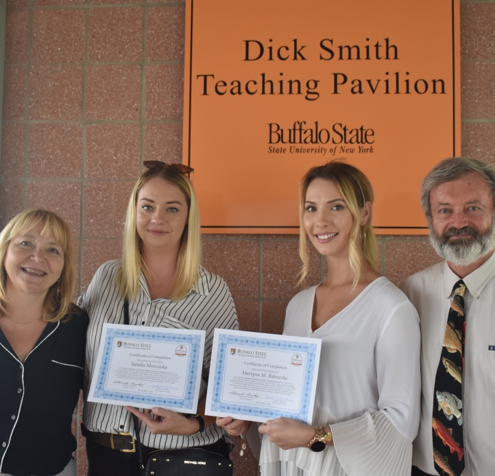 Four people posing for a picture by a wall. The two in the center are holding certificates. A sign behind them says "Dick Smith Teach Pavilion, Buffalo State."