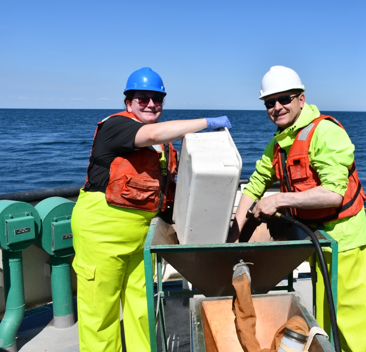 Two smiling people in life jackets and hard hats work at a basin on the deck of a large boat. One person dumps a tub into the basin while the other uses a hose to wash the tub
