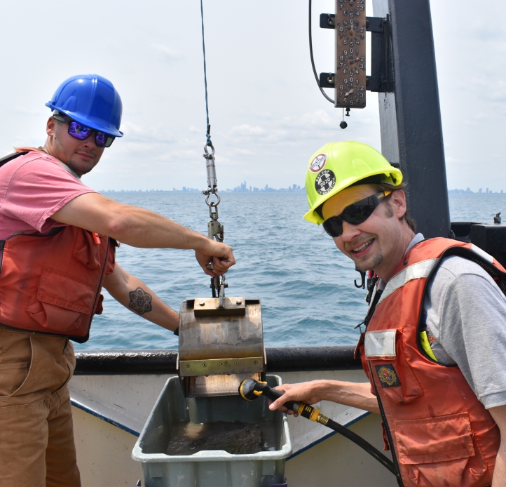 Two people working on a boat on a lake, wearing hardhats and life jackets. One holds a metal scoop with mud over a tub while the other washes it out.