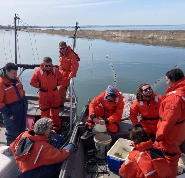 A group of people on two work boats cluster around a cooler with fish. Everyone is wearing coldwater safety gear.