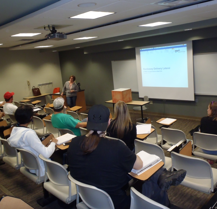 A lecturer stands before students seated in a lecture hall. A projector screen has a power point slide on it.