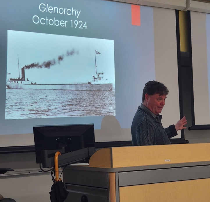 A person lecturing at the front of a room standing in front of a projector screen that says "Glenorchy October 1924" with a black-and-white photo of a freighter ship