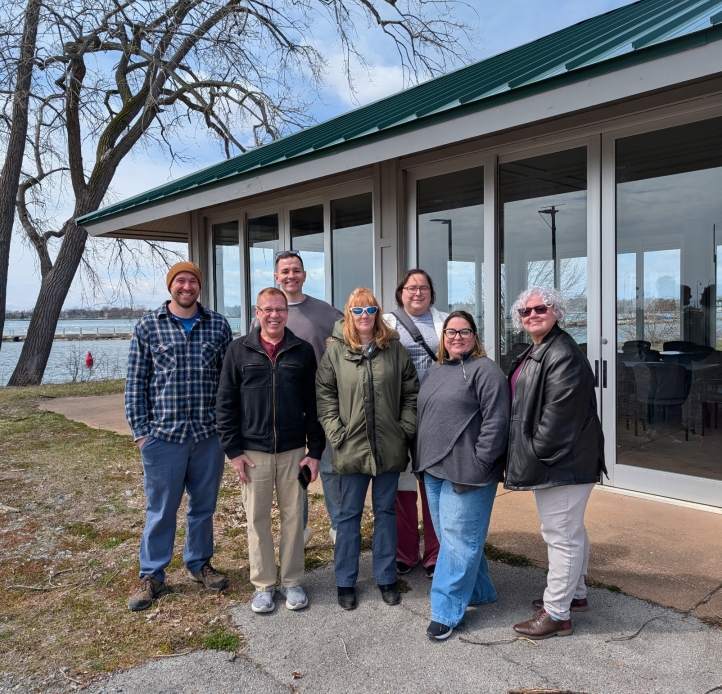 Seven people pose for a picture outside a small building with floor-to-ceiling windows at the waterfront.