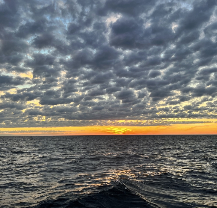 Sunset on a large body of water. A thin band of orange rests between gray dappled clouds and steely waves.