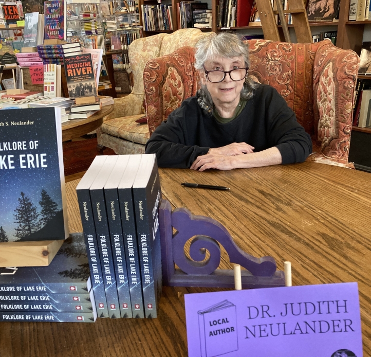 A person sits in an armchair behind a table with a stack of books "Folklore of Lake Erie" in a bookstore with a pen beside them for signing; tag "Local author Dr. Judith Neulander"