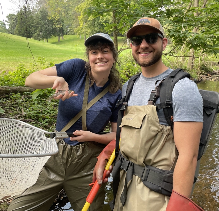 Two people wearing waders stand smiling in a stream. One has a backpack unit, a pole, and thick rubber gloves, while the other holds a net and a small fish.