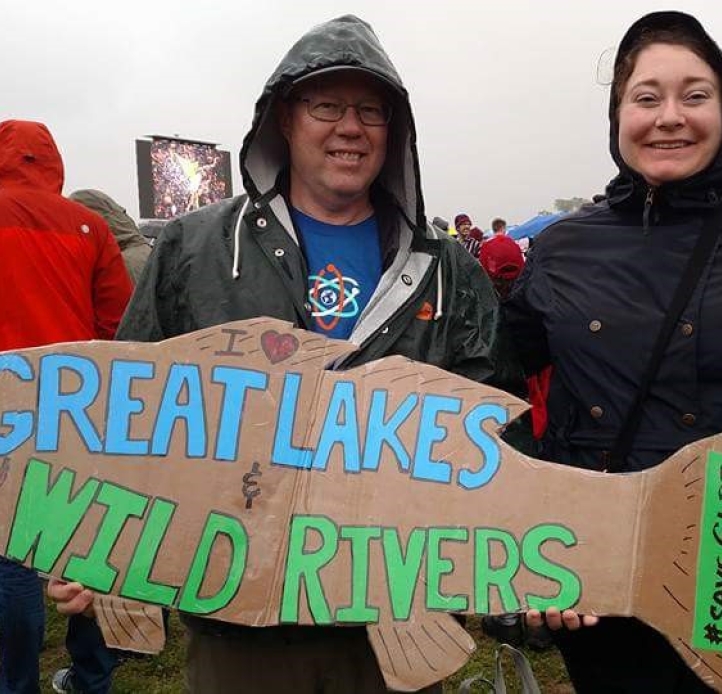 Two people in rain coats pose for a picture amidst a crowd of people. One is holding a sign shaped like a fish that says "I [heart] Great Lakes & Wild River #SaveGLRI"