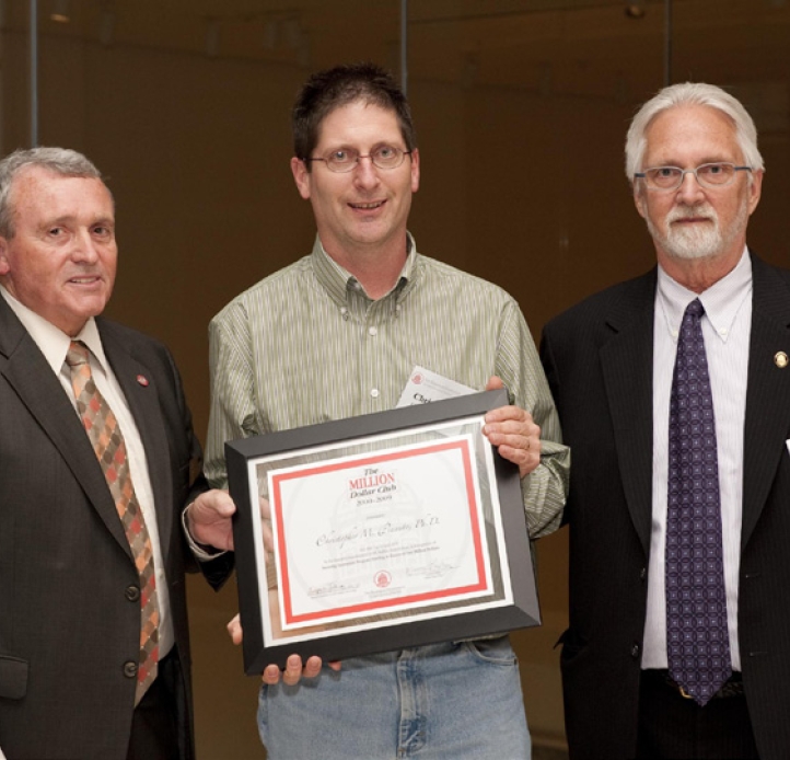 Two people in suits flank a person in a button-down shirt holding a framed certificate as they pose together