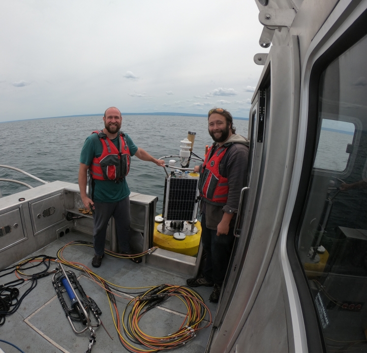 Two people in life jackets stand on the aft deck of a boat, holding a buoy that floats alongside the boat on an overcast day.