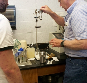 A person takes a metal lid with two rods and a tiny metal cup and prepares to set it on a stand. Nearby is a lab instrument and a large metal cup that the lid fits on. Someone watches.