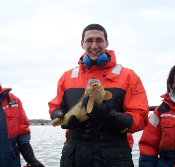Three people wearing cold water safety gear by the water smile and laugh. The person in the middle holds up a carp