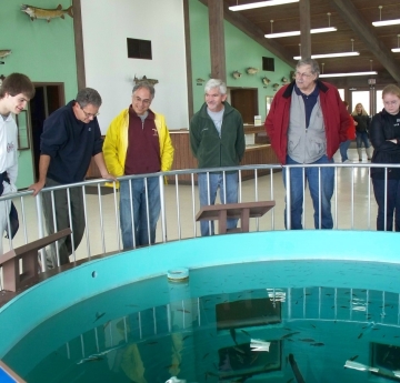 A group of people stand around a large round aquarium in the floor, leaning on the railing. The wall behind them has mounted fish.